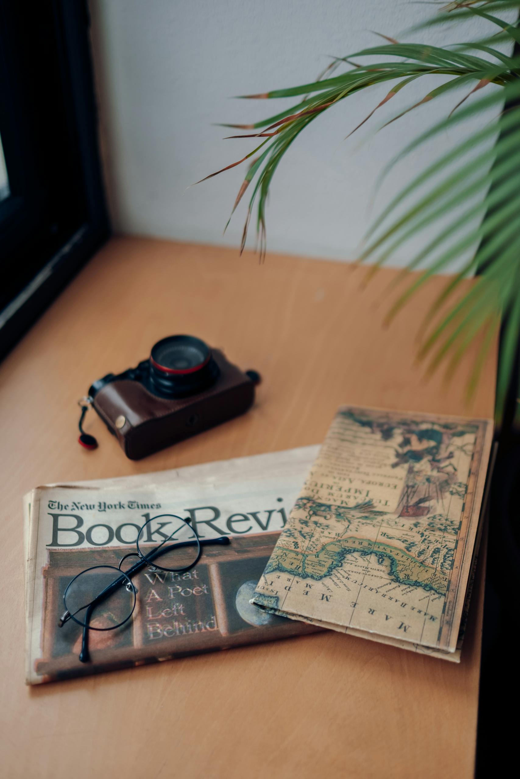 Vintage camera, book review, and antique map placed on a wooden table next to glasses and plant.