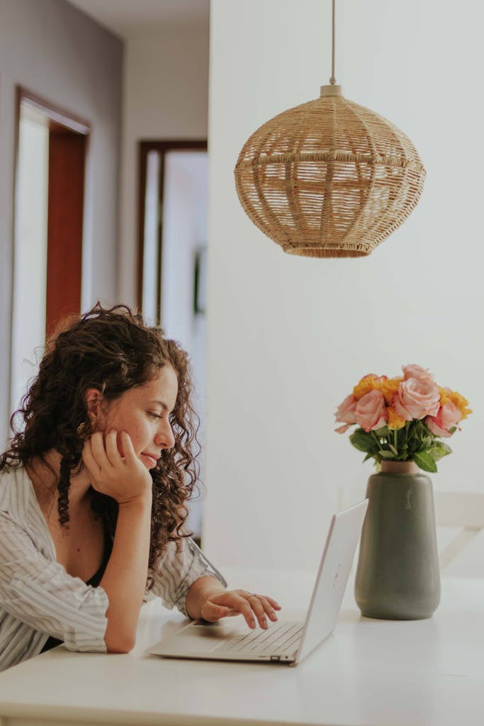 A woman uses a laptop at home with flowers and a woven lamp, creating a cozy work environment.