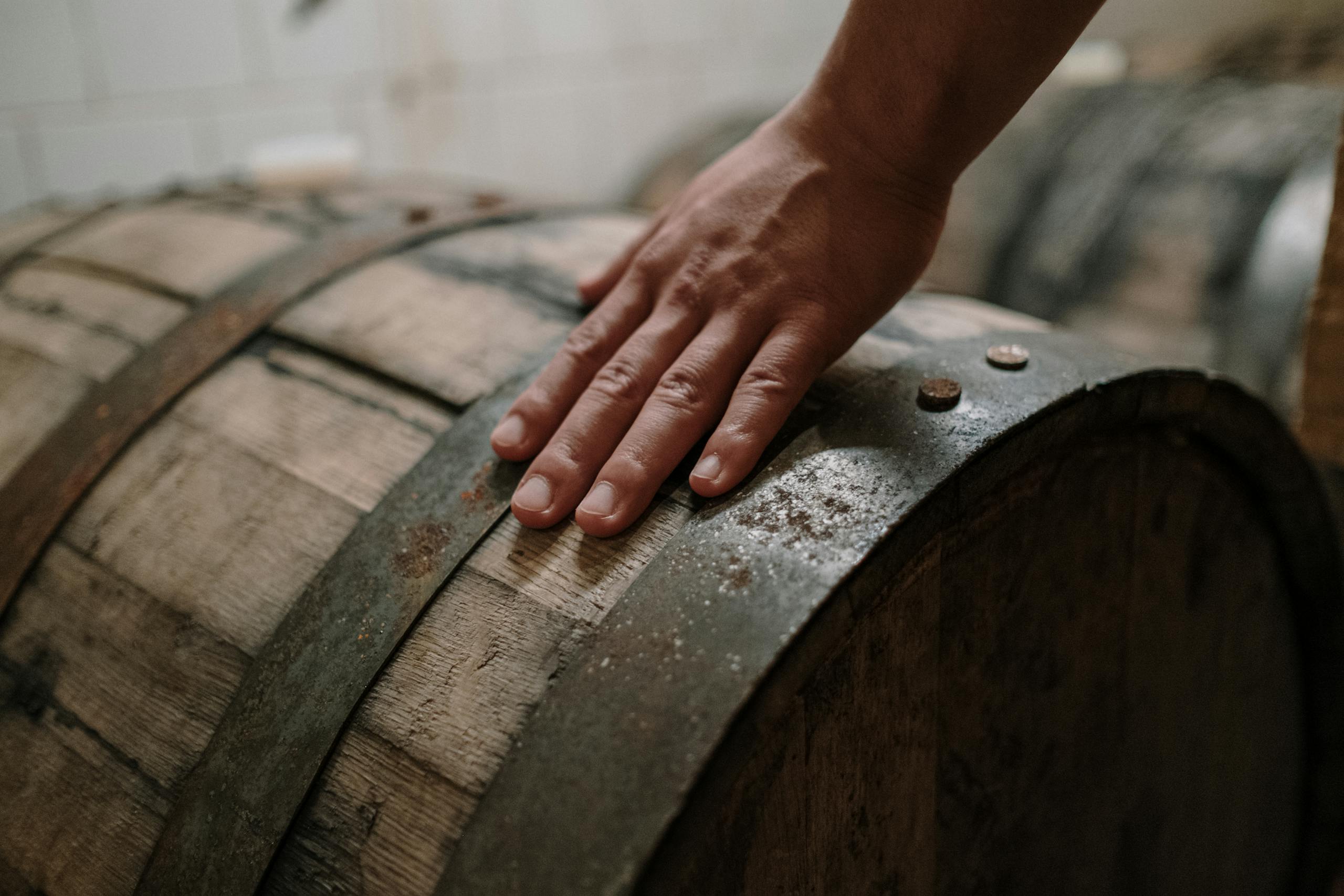 Close-up of a hand resting on a rustic wooden barrel in a brewing environment.